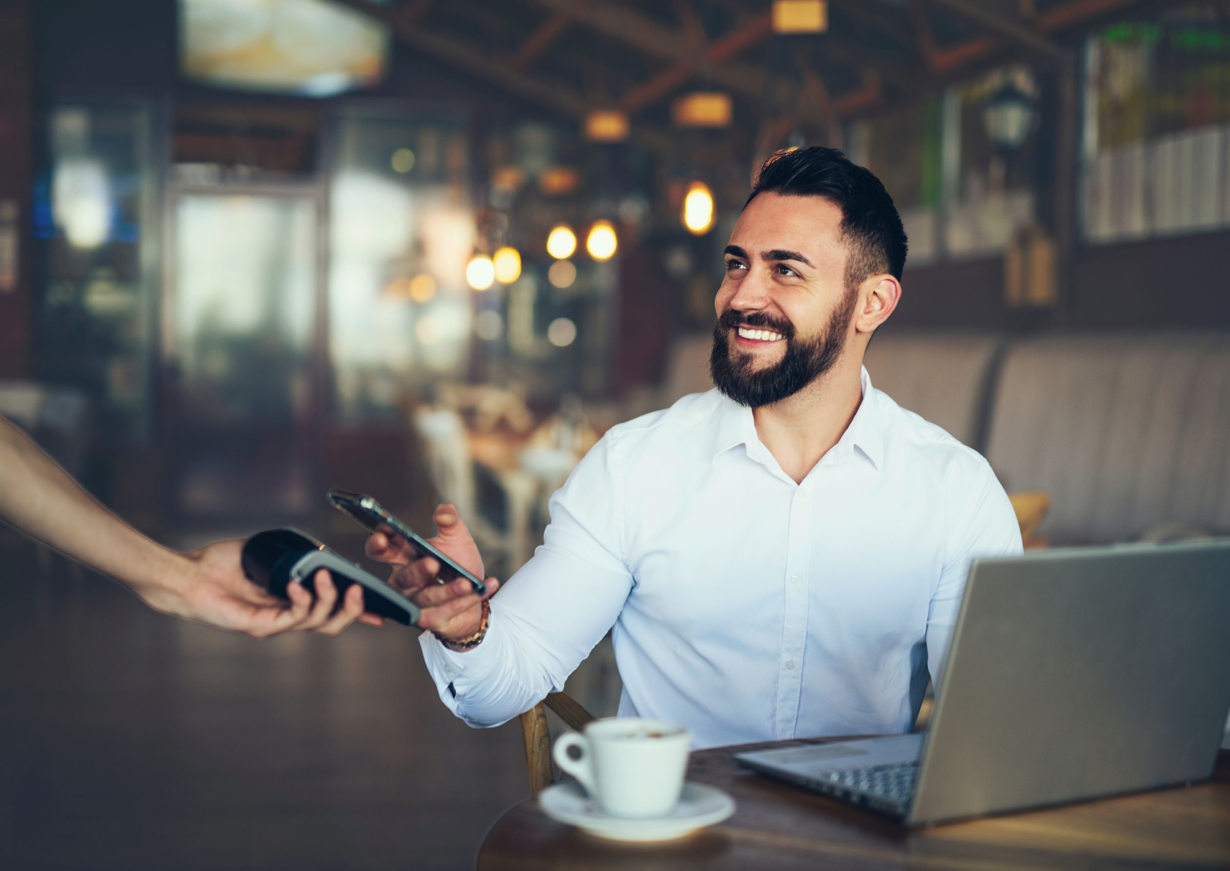 Lächelnder Mann in weißem Hemd sitzt mit einem Laptop und einer Kaffeetasse an einem Tisch in einem Café und hält sein Smartphone an ein mobiles Kartenlesegerät, um kontaktlos zu bezahlen.